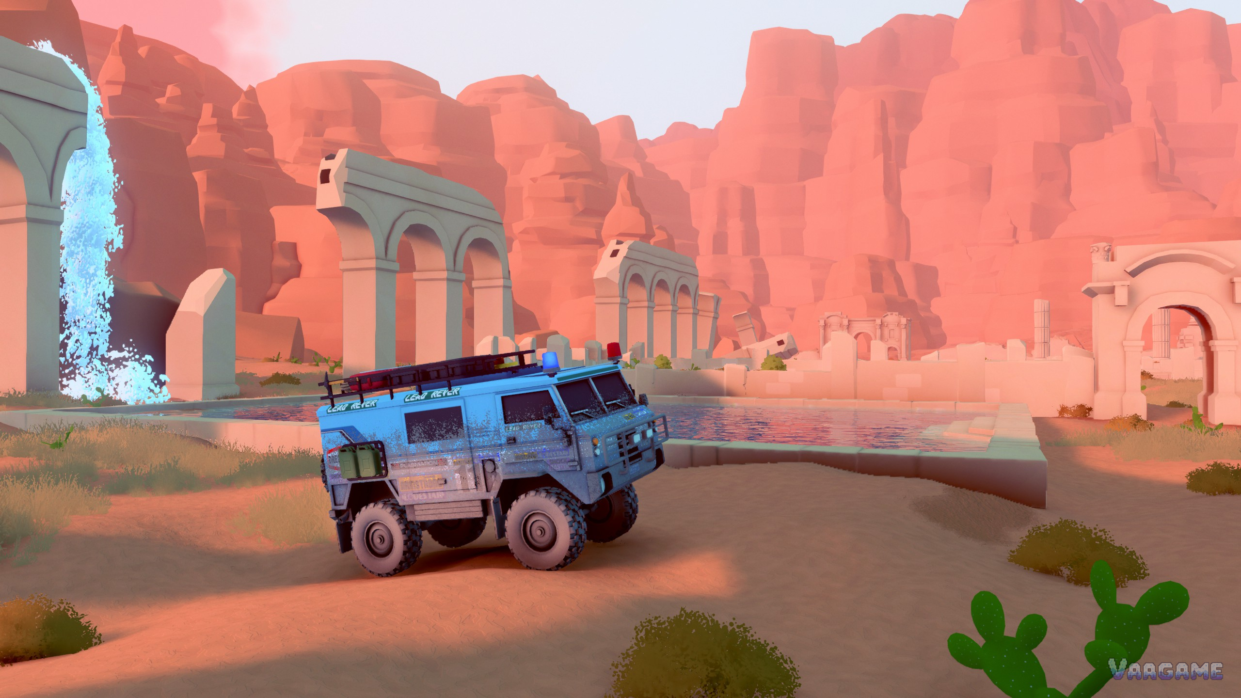 A blue six-wheeled expedition truck before ruined aqueduct arches and a waterfall pool